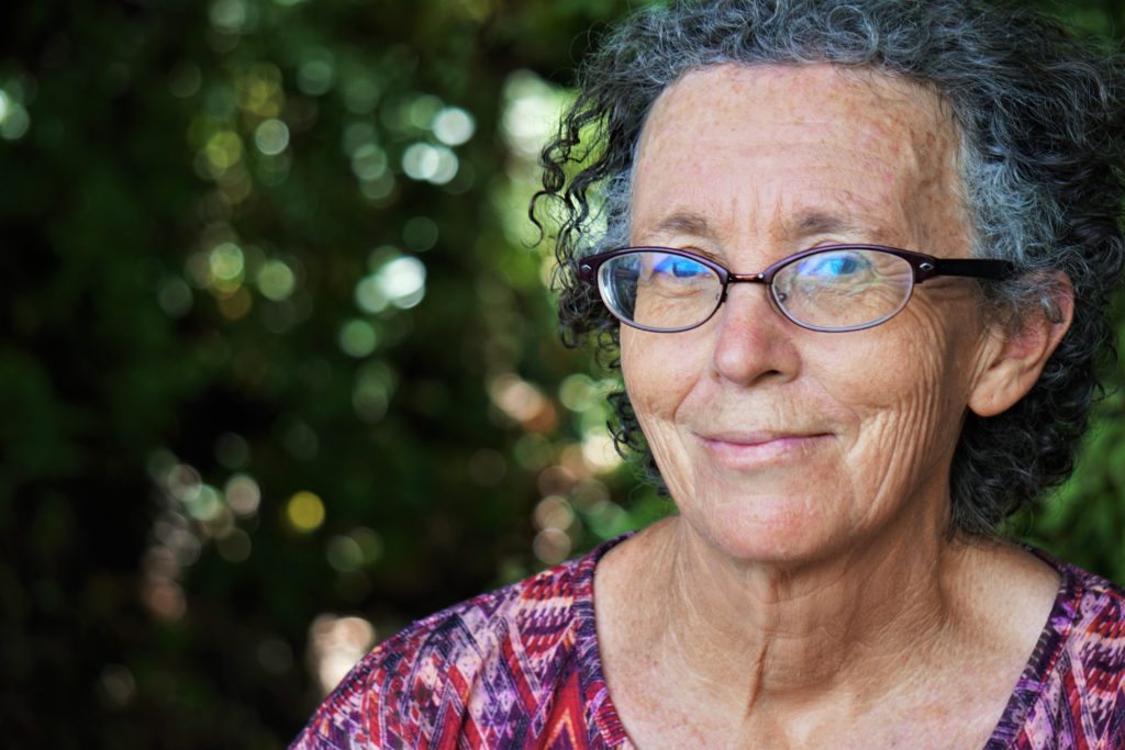 CLR vs RLE; There is an older woman seen from the shoulders up, wearing a pink shirt and glasses smiling gently at the camera. There are trees and greenery making up the background