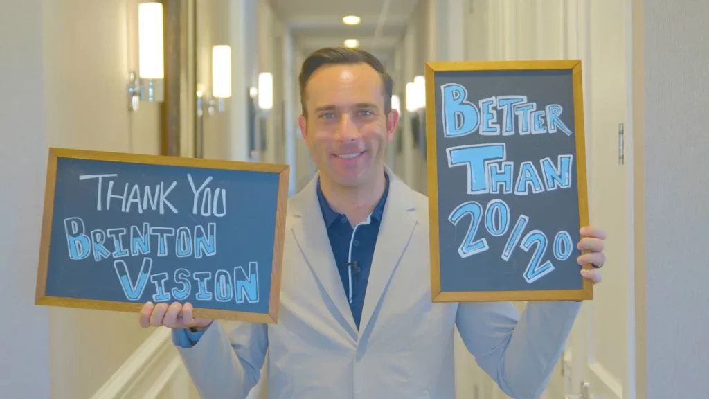 photo of dave, a white man in a blue button up and white lab coat from the chest up. He is standing in a well lit hallway holding two chalkboard signs. The one in his right hand says "Thank you Brinton Vision", the one in his left hand says "Better than 20/20"